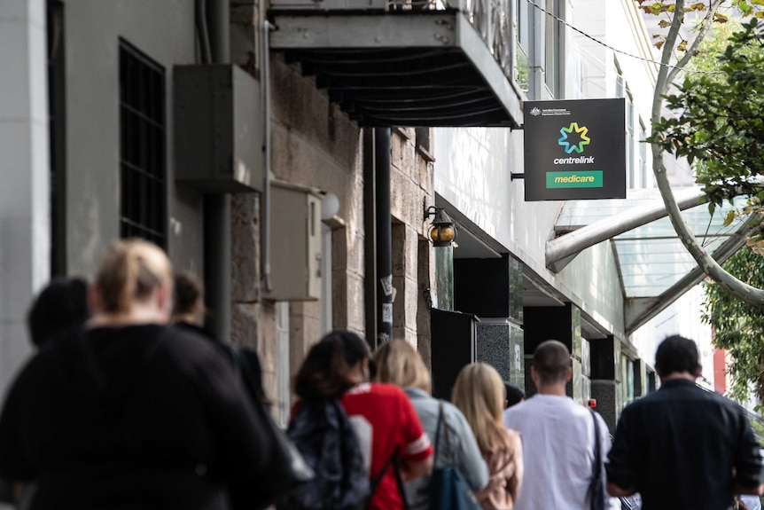 A line of people photographed from behind in front of a sign that reads Centrelink.