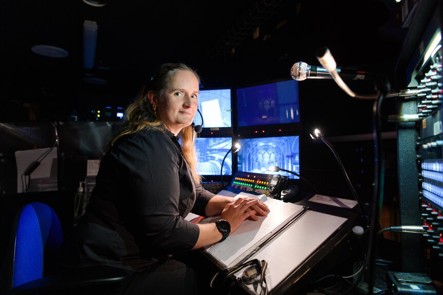 Woman with long blonde hair wears black clothes and headset while sitting at stage management desk in theatre wing.