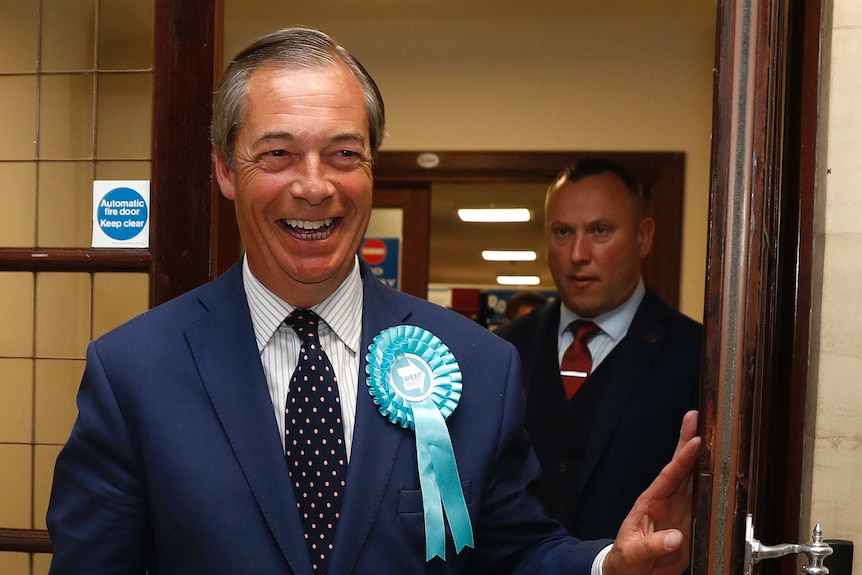 Nigel Farage walks through a door with a Brexit ribbon on his jacket lapel.
