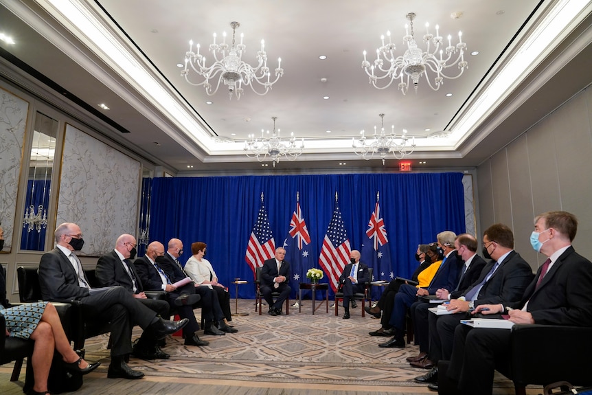 Two men in suits sit beside each other and look towards two rows of people seated opposite each other.