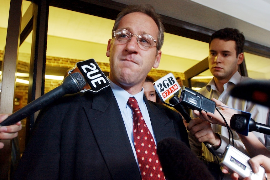 A man wearing glasses is clearly close to tears as he faces a line of microphones