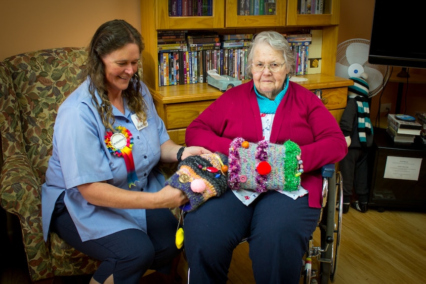 Heather Robertson sitting next to Grace Potter, who is in a wheelchair, in a nursing home room and and they hold fiddle muffs.