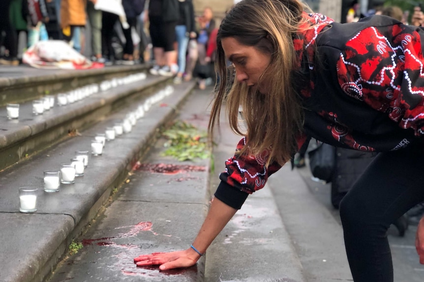 A woman with blood-like red fluid on her hand, puts her palm on a step outside the Victorian Parliament.