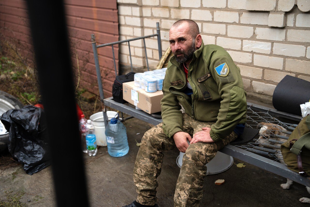 A man in a military uniform sitting on a bed frame, outdoors, looking on