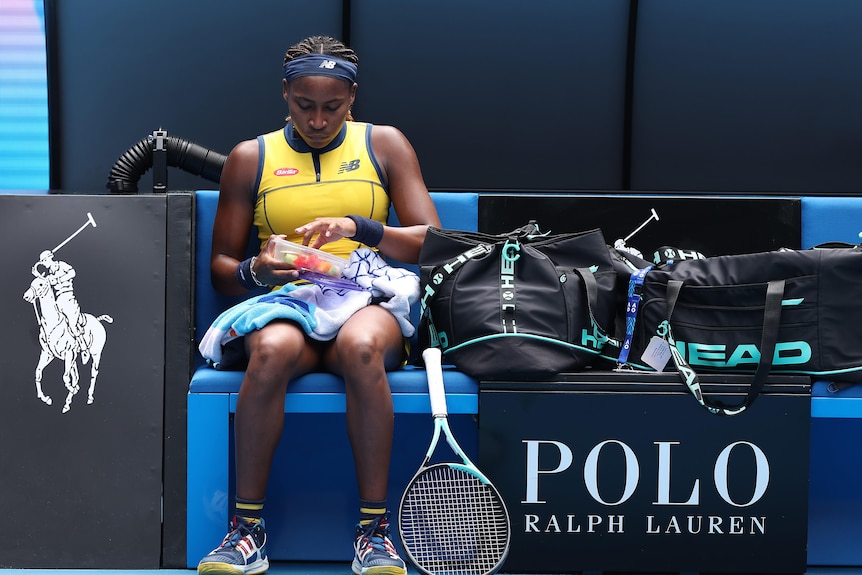 Female tennis player Coco Gauff sitting on her courtside bench, eating fruit