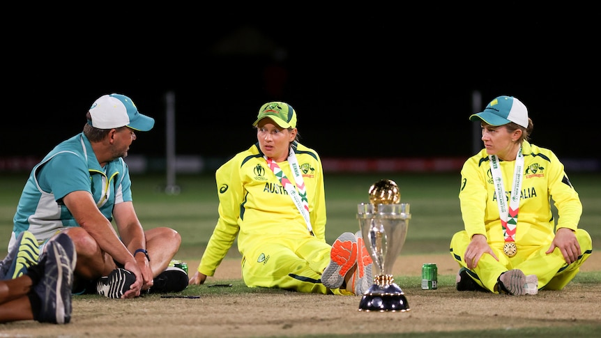 Matthew Mott, Meg Lanning and Rachael Haynes all sit on the ground next to the World Cup trophy