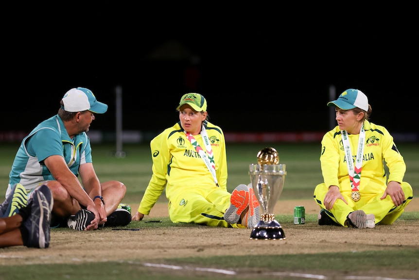 Matthew Mott, Meg Lanning and Rachael Haynes all sit on the ground next to the World Cup trophy