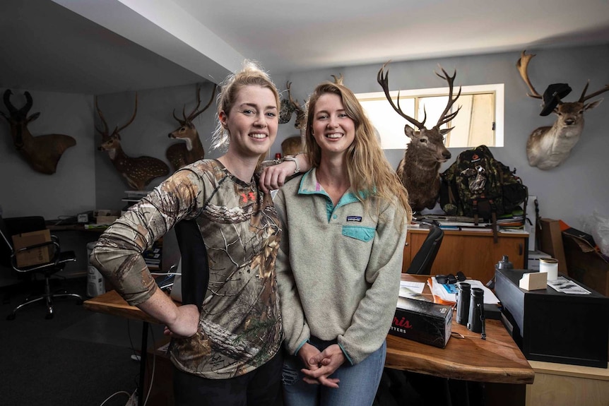 Two women stand in front of a desk with a wall of trophy heads behind them.