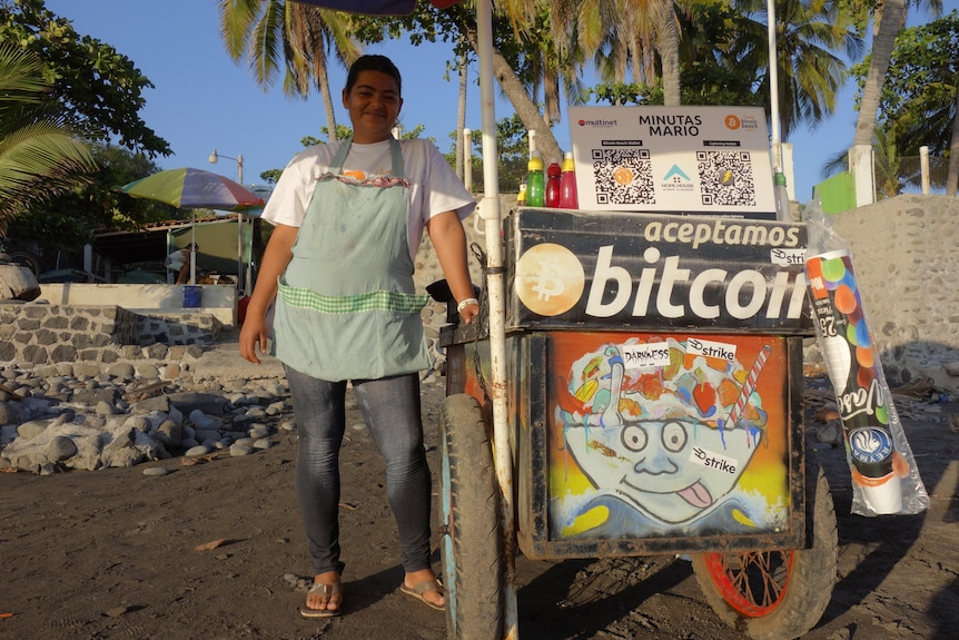 A man stands next to a food cart decorated with posters saying 'aceptamos bitcoin'