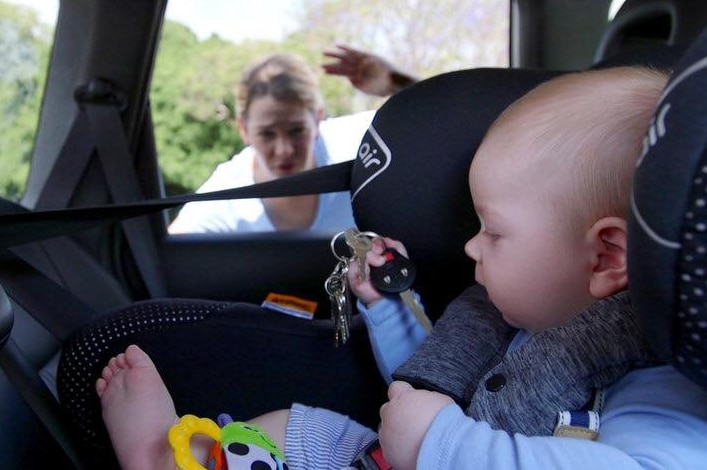 A baby sits in a car seat holding car keys, while the mother looks on from outside the car.