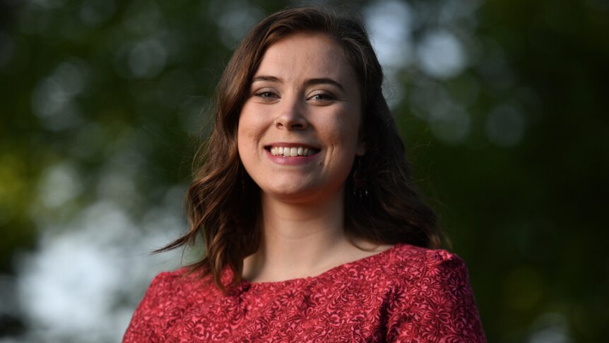 Madeline Diamond smiling in a red dress with trees out of focus in the background