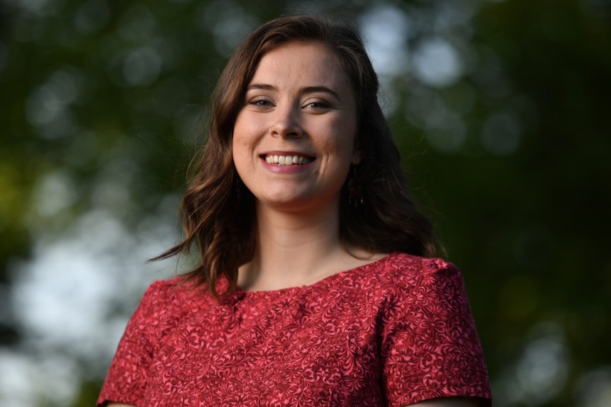 Madeline Diamond smiling in a red dress with trees out of focus in the background