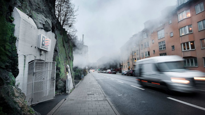 A white concrete bunker is built into the side of a granite cliff with an urban scene on the opposite side of the street.