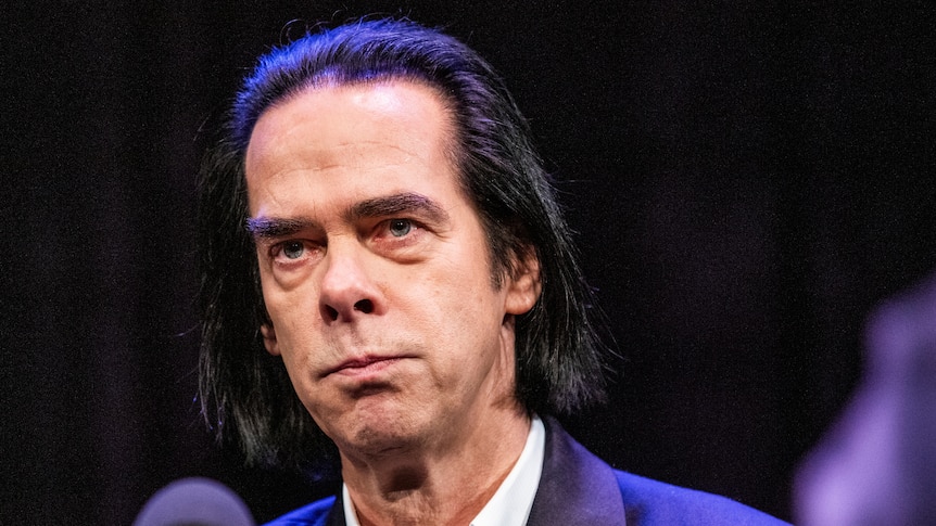 Nick Cave, a middle-aged white man with long dark hair and wearing a suit, speaks into a microphone