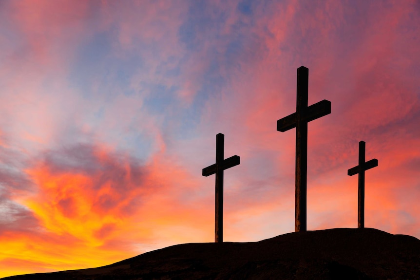 The silhouette of three crucifix stand atop a mountain, against a backdrop of a sunset.