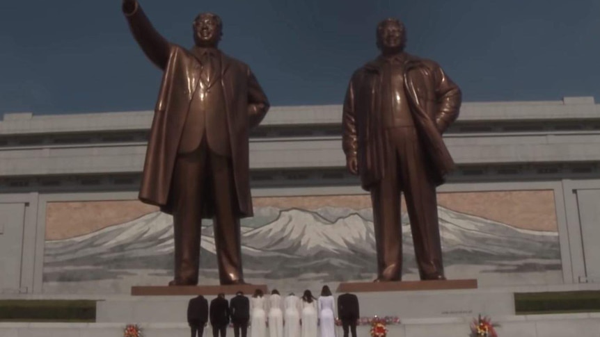 A group in formal attire bow before two large bronze statues