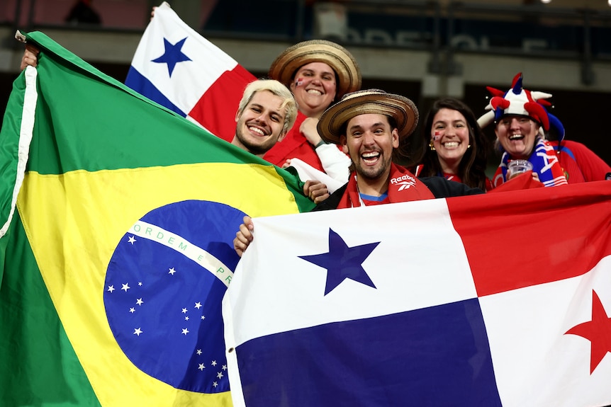 Brazilian and Panamanian fans smile at the camera as they hoild their national flags in the stands before a game.