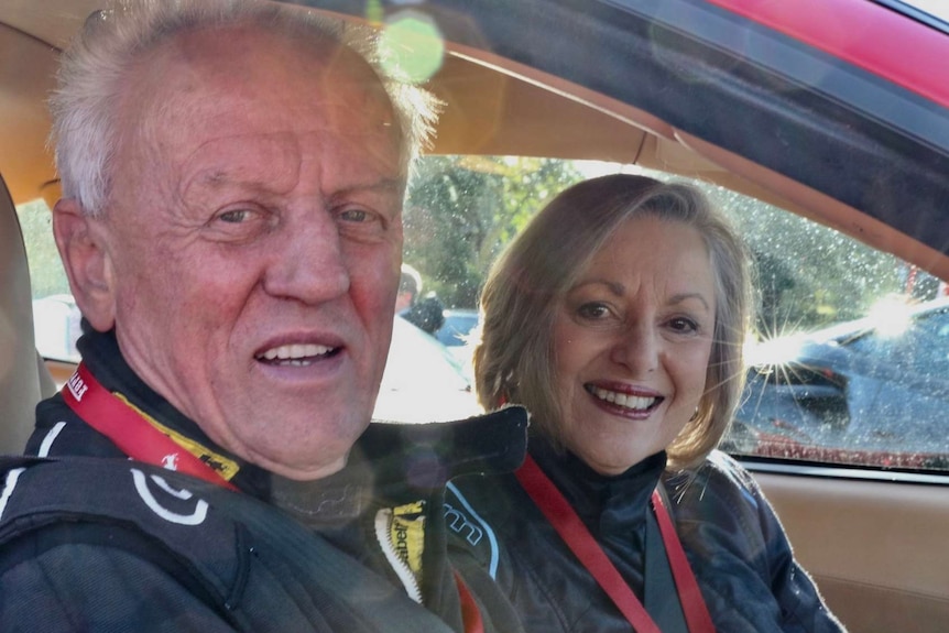 Conrad Whitlock sits in a car in a race suit, while Mandy smiles from the seat next to him.