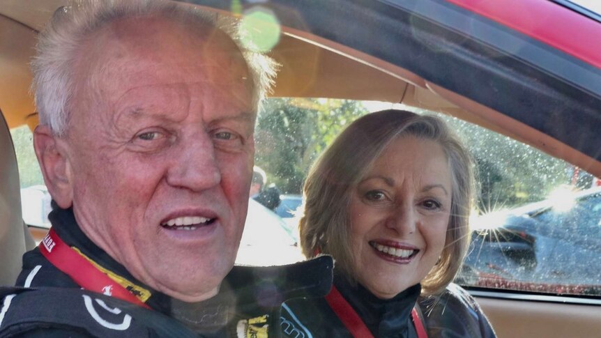 Conrad Whitlock sits in a car in a race suit, while Mandy smiles from the seat next to him.