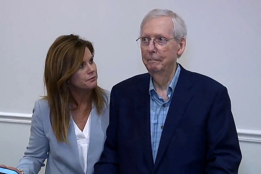 An old man wearing a suit and glasses staring off camera in a stunned look with a woman beside him looking at him