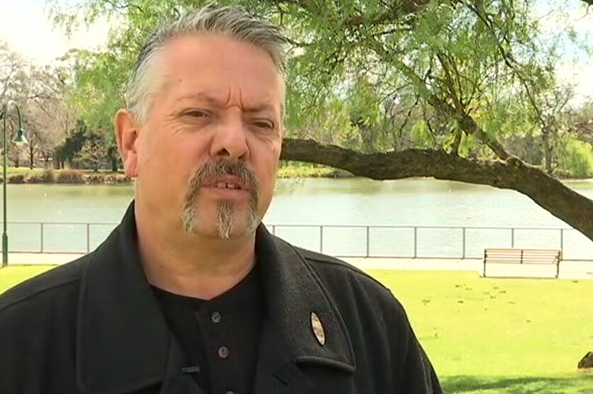 Wearing a dark jacket, Rodney Carter stands in front of a tree and a body of water as he is interviewed for television.