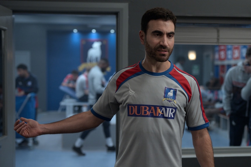 A 40-something man in a soccer jersey looks angry, gesturing to his right, in a team locker room 