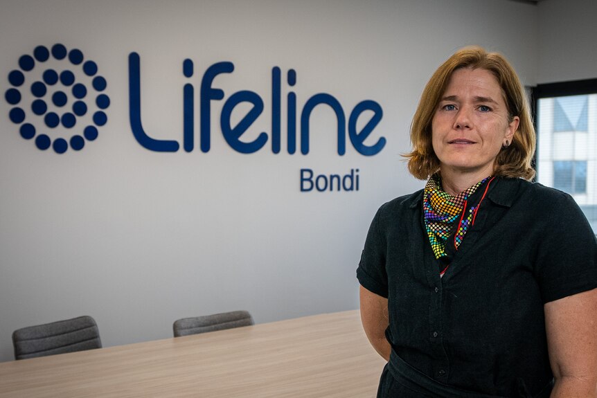 A middle aged woman wearing a black polo shirt and colourful scarf stands in front of a Lifeline logo in a meeting room.