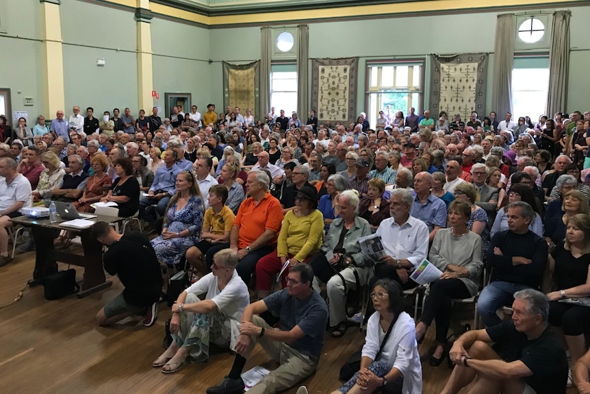 A large crown of people pack inside a town hall building.