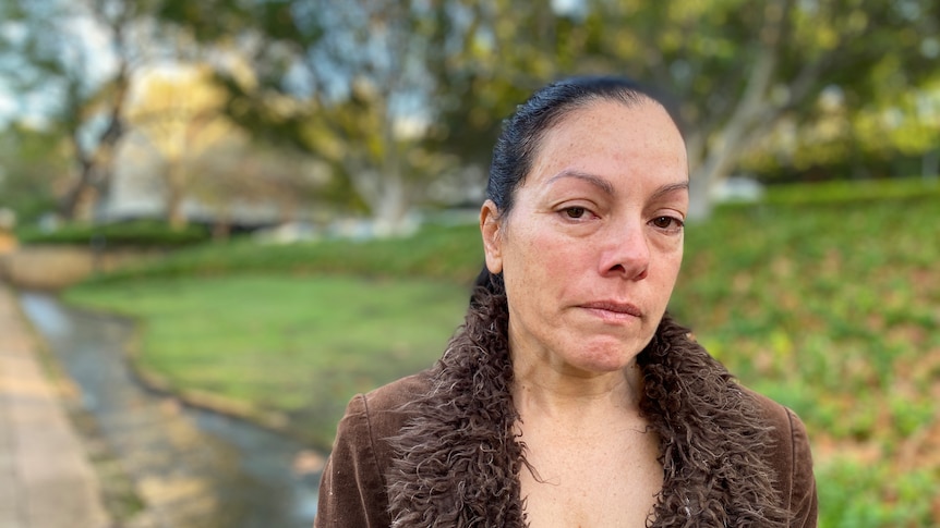 A woman in a brown coat looks at the camera - she is standing on a footpath in East Perth with a grassy bank behind her