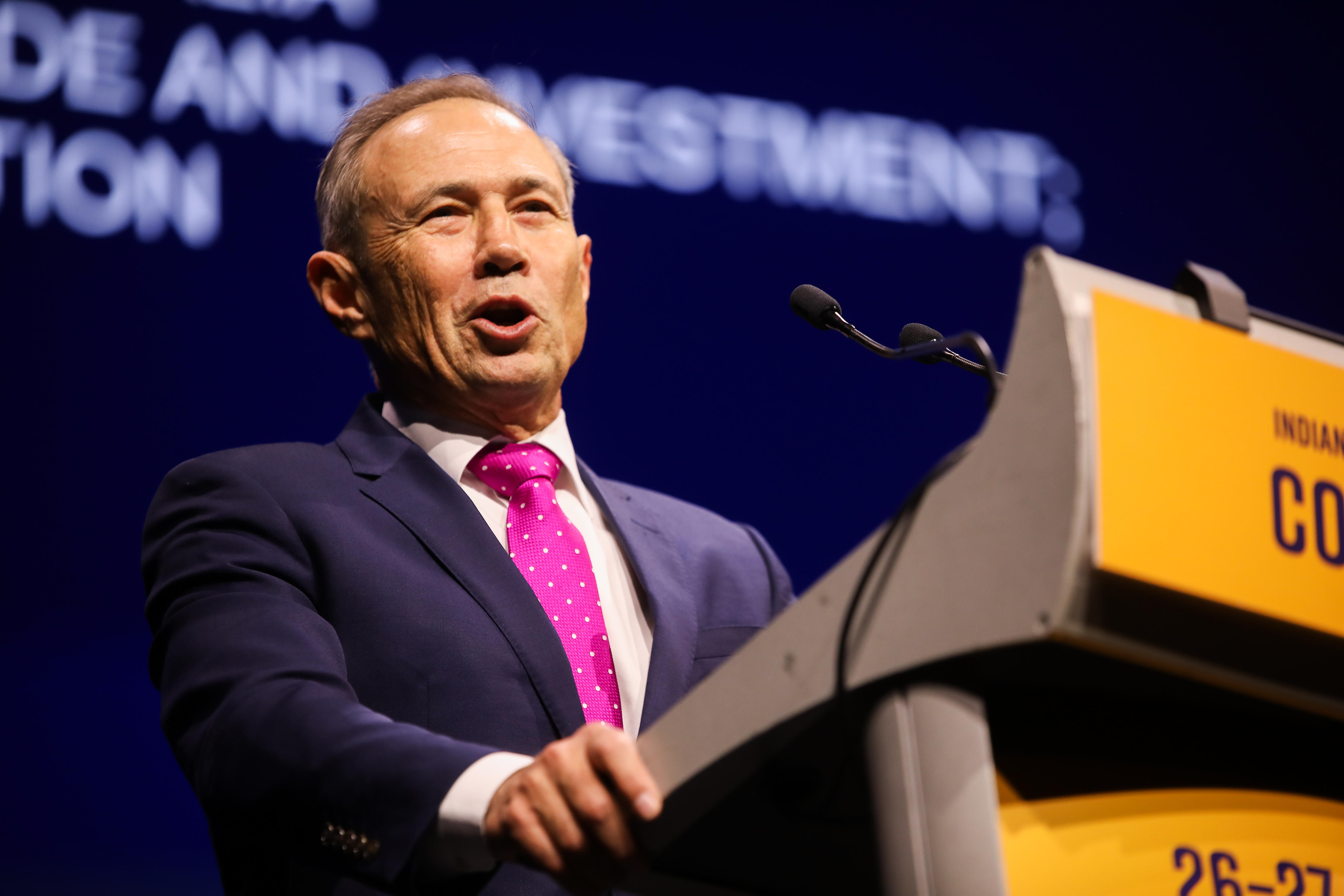 A mid-shot of WA Premier Roger Cook giving a speech at an orange dais on stage wearing a suit and tie, against a black backdrop.