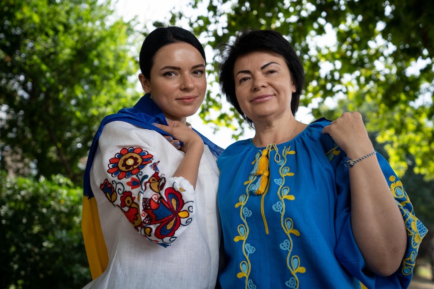 Две женщины стоят близко друг к другу, одетые в вышитые цветочные топы (одна сине-желтая), вокруг них накинут украинский флаг.