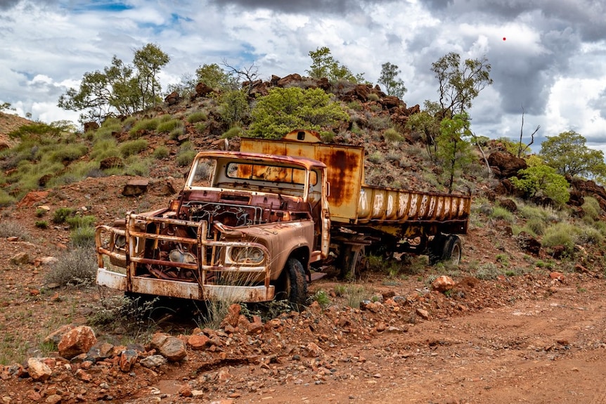 A vintage truck has rusted away about 1 metre from the red dirt road