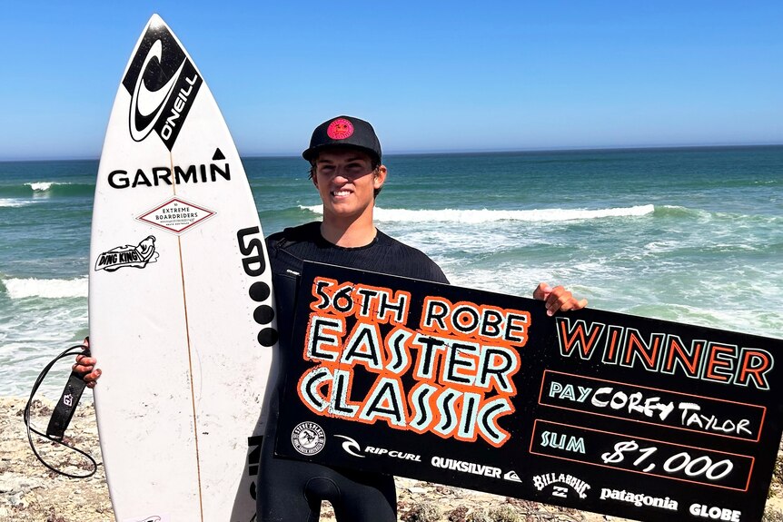 A teenager holds a surfboard and a winner's cheque