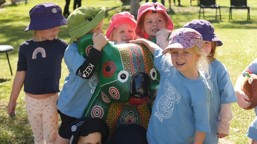 Preschool children in colourful hats stand with arms around a koala statue painting in green and Aboriginal designs.