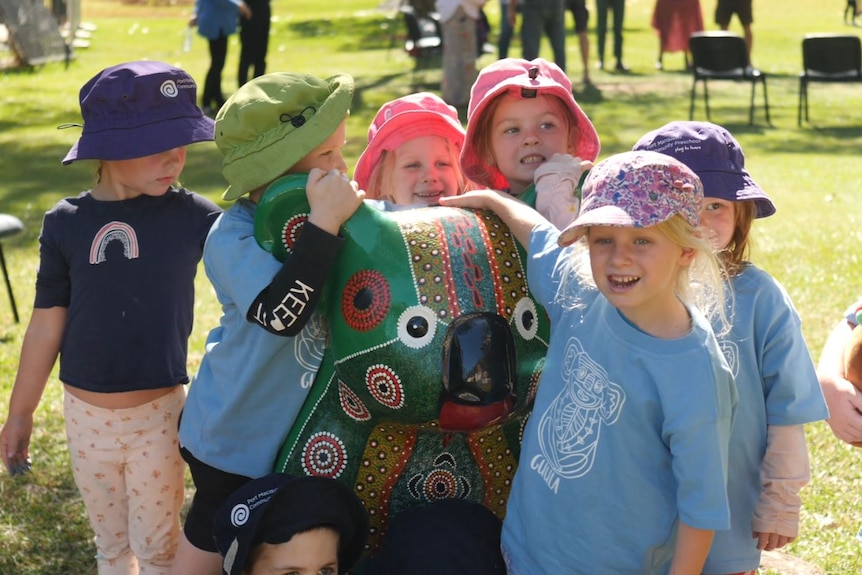 Preschool children in colourful hats stand with arms around a koala statue painting in green and Aboriginal designs.