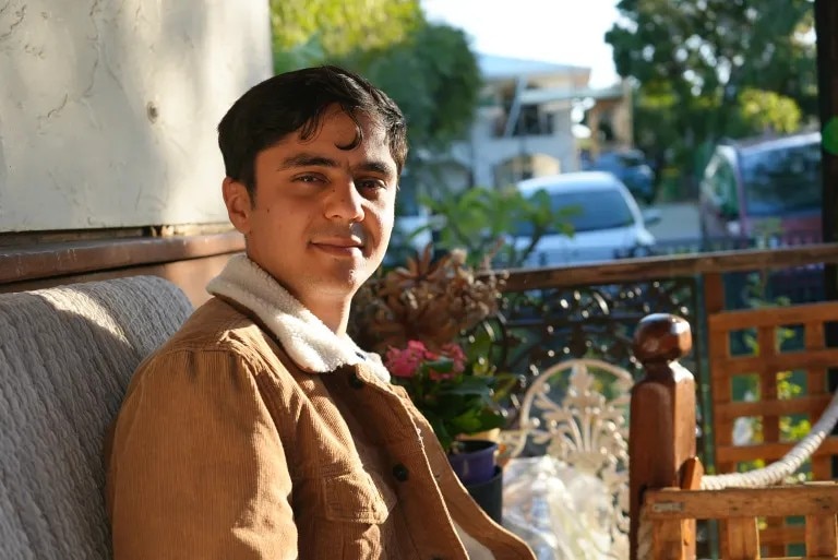A man with dark hair wearing a camel coloured sherpa jacket looking at the camera whilst sat on an outdoor couch