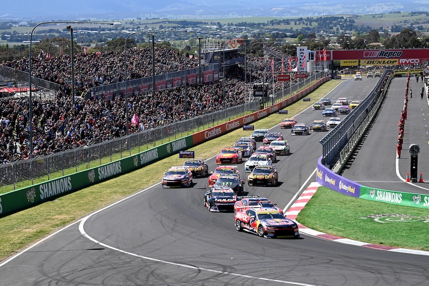 An ariel view of several race cars at the first corner of a race, in front of fans.