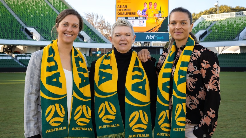 Three women wearing green and yellow Australia scarves stand in an empty stadium to pose for a photo