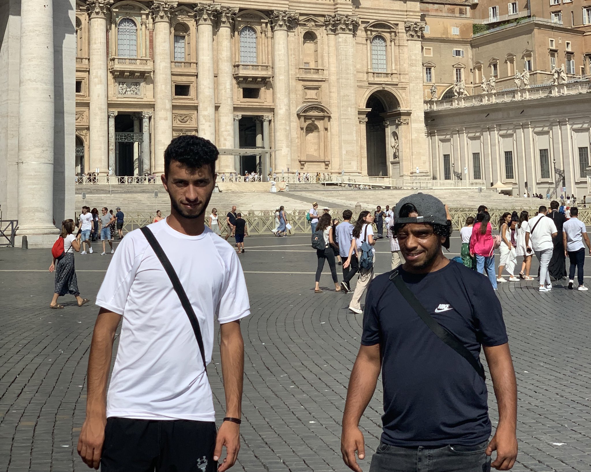 Two men wearing t-shirts stand side-by-side in a town square with large sandstone buildings behind them