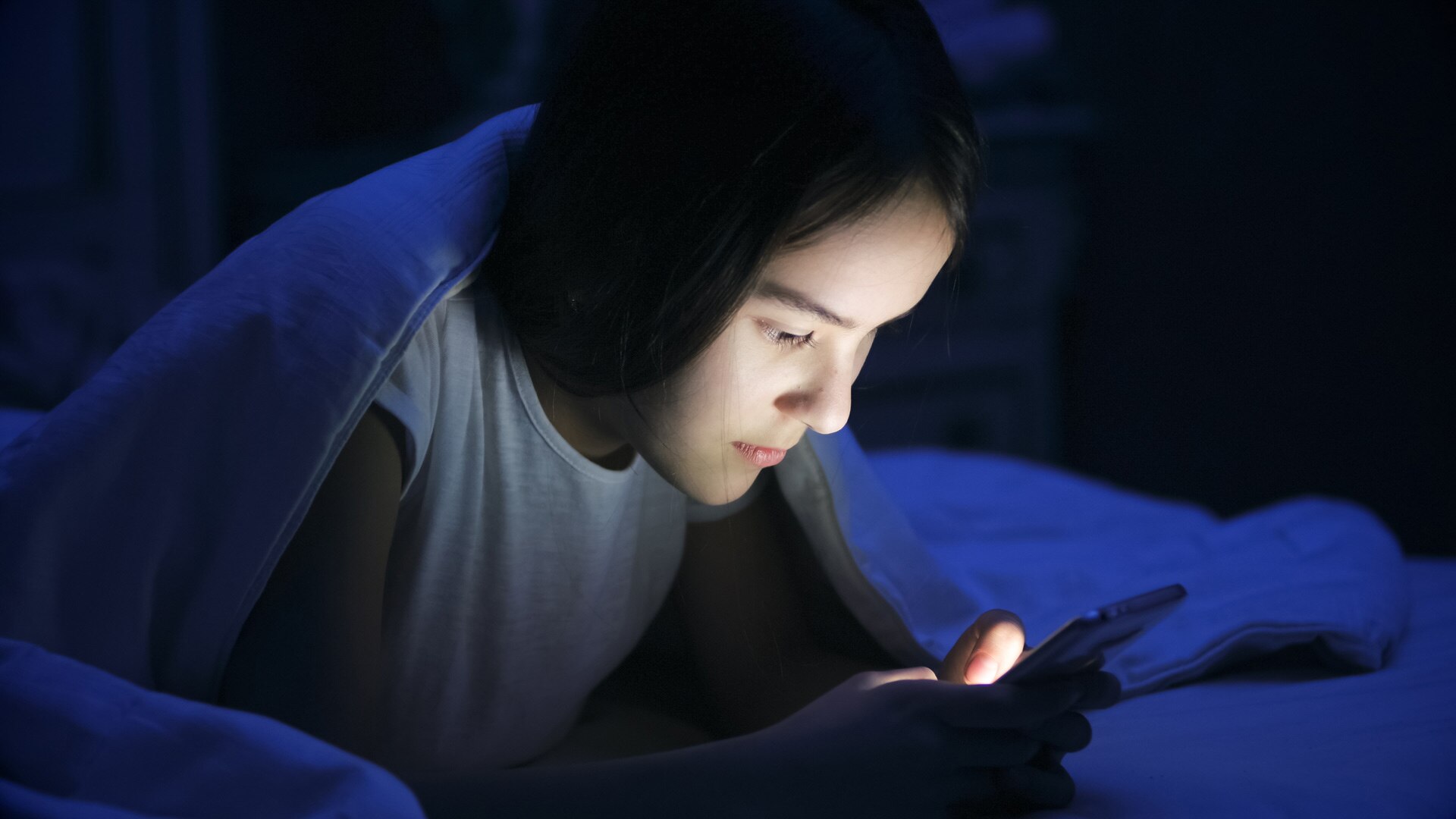 A teenage girl supports herself on her elbows in bed and uses her phone in the dark.