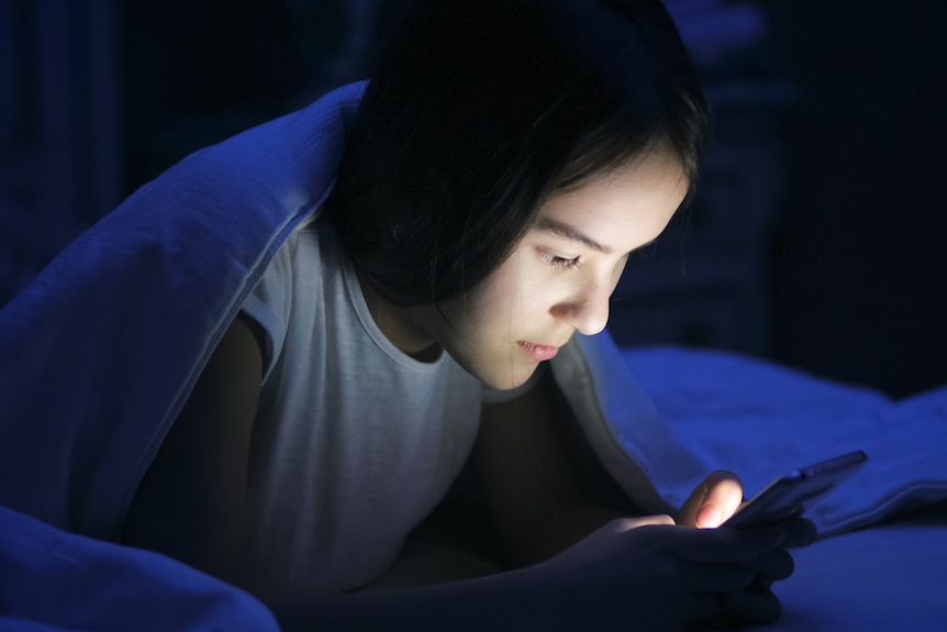 A teenage girl supports herself on her elbows in bed and uses her phone in the dark.