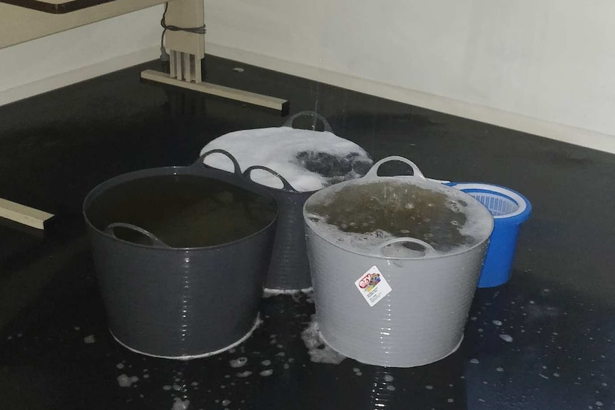 A room with a desk has four buckets on the ground filled with water from flooding coming through the roof.