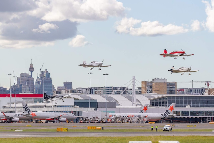Three small planes in the air over a runway at Sydney Airport with jets in the background.