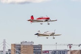 Three small planes in the air over a runway at Sydney Airport with jets in the background.