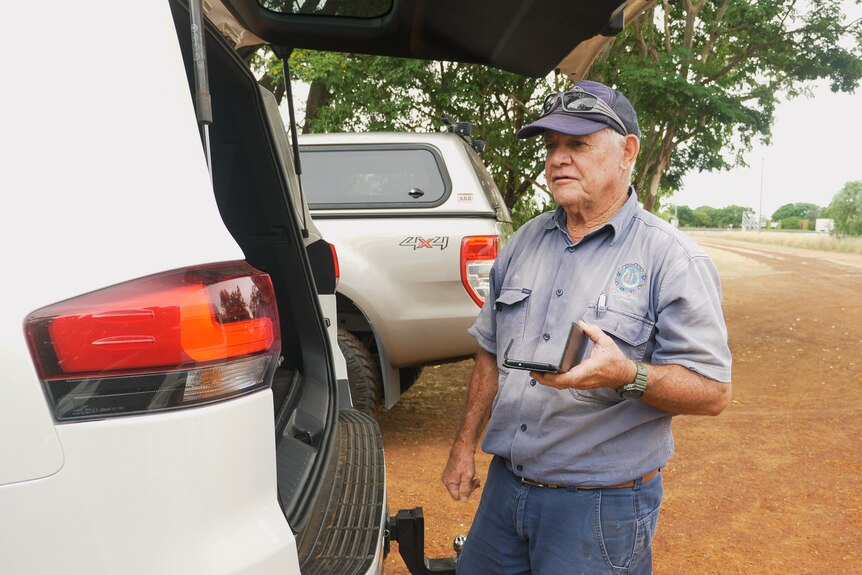 Ron Waters stands near a white car holding a phone, St George, Queensland, March 2024