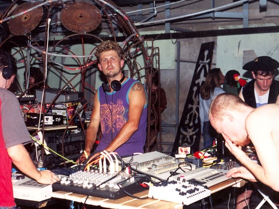 Four men play electronic musical instruments in front of a dome made from rusting welded metal.