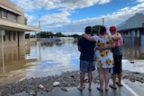Three people stand with arms around each other, looking over floodwaters around a town's main street.