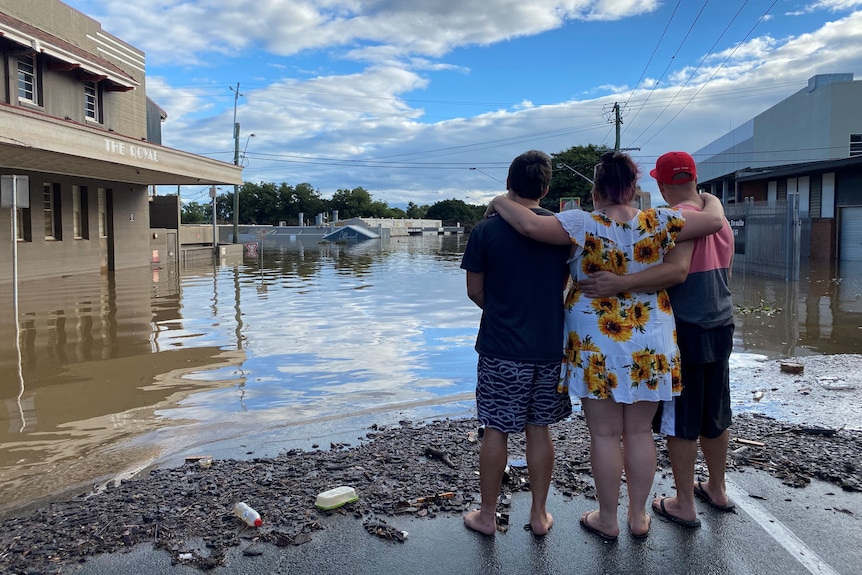 Tres personas se abrazan y miran por encima de las inundaciones en la calle principal de un pueblo.