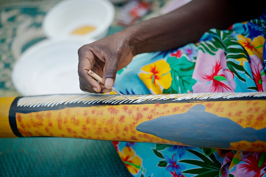 The hand of an Indigenous woman can be seen delicately painting a wooden yidaki.