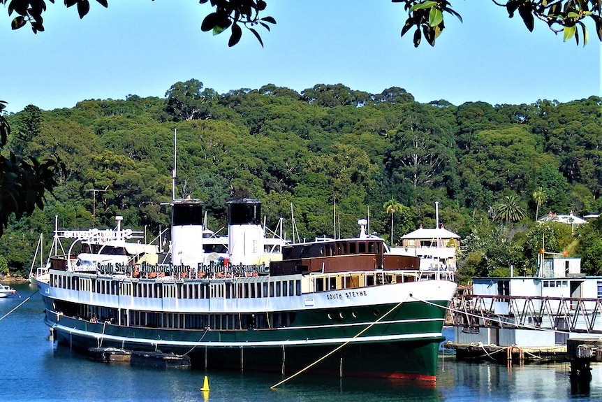 A large steam ferry moored at a bay with trees in the background.